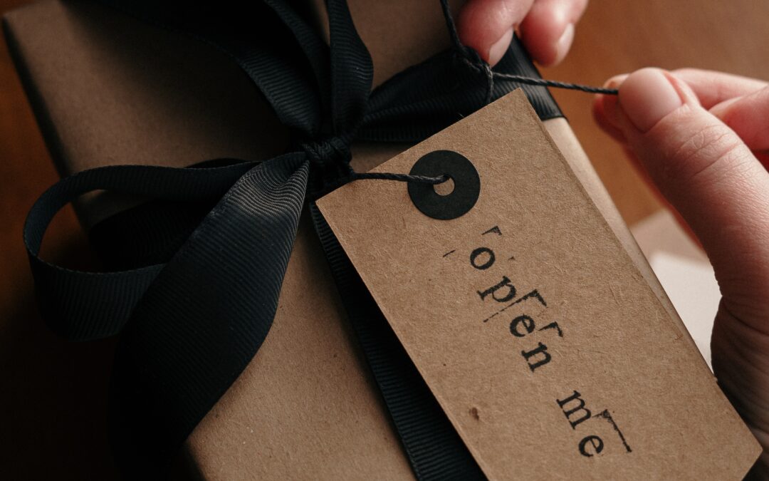 Δύο χέρια τυλίγουν με μπεζ χαρτί ένα δώρο με έναν μαύρο φιόγκο στον οποίο υπάρχει ένα κομμάτι χαρτί που γράφει "open me".